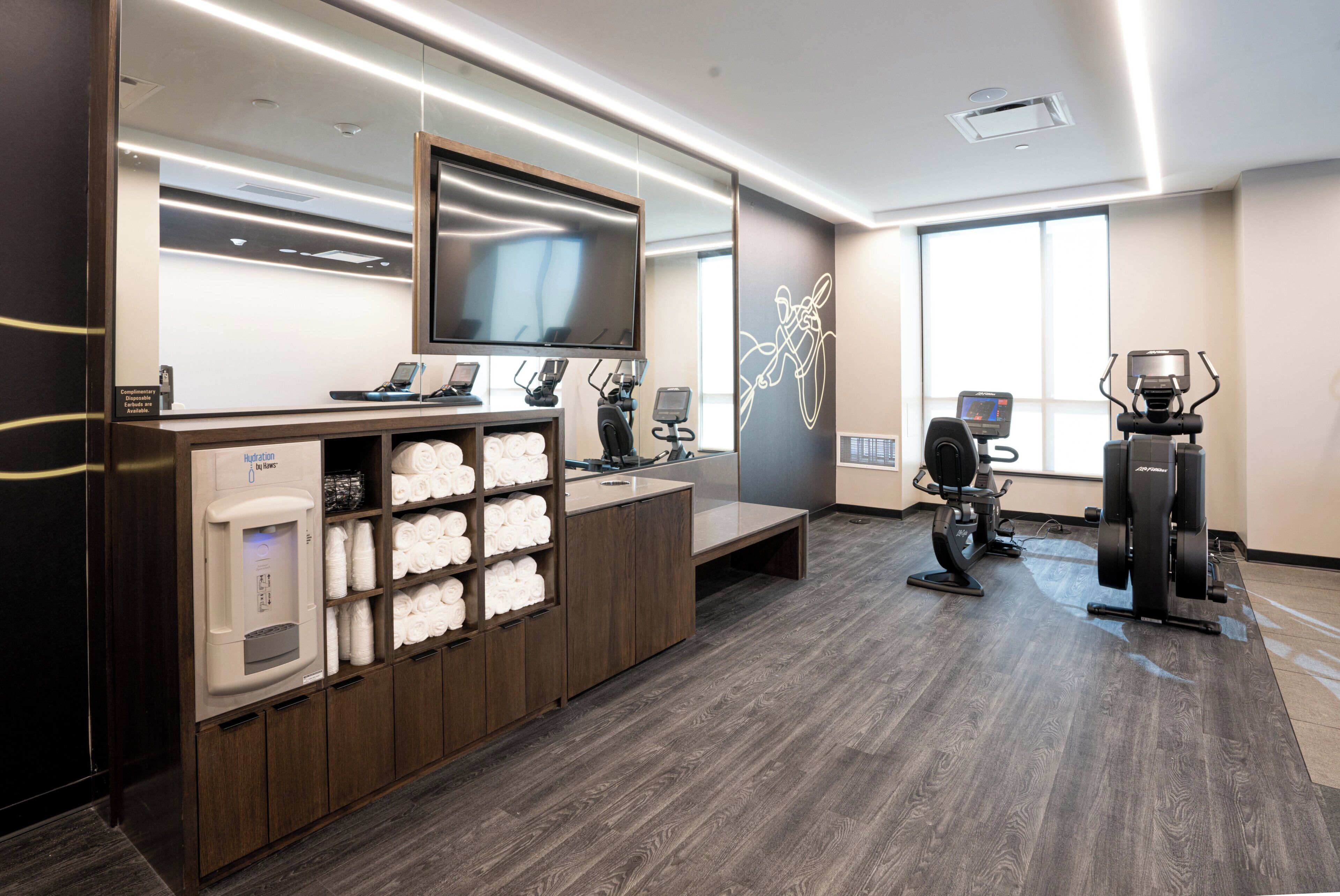 A modern spa room with a large mirror, a television, a water fountain, exercise equipment, and neatly arranged towels.