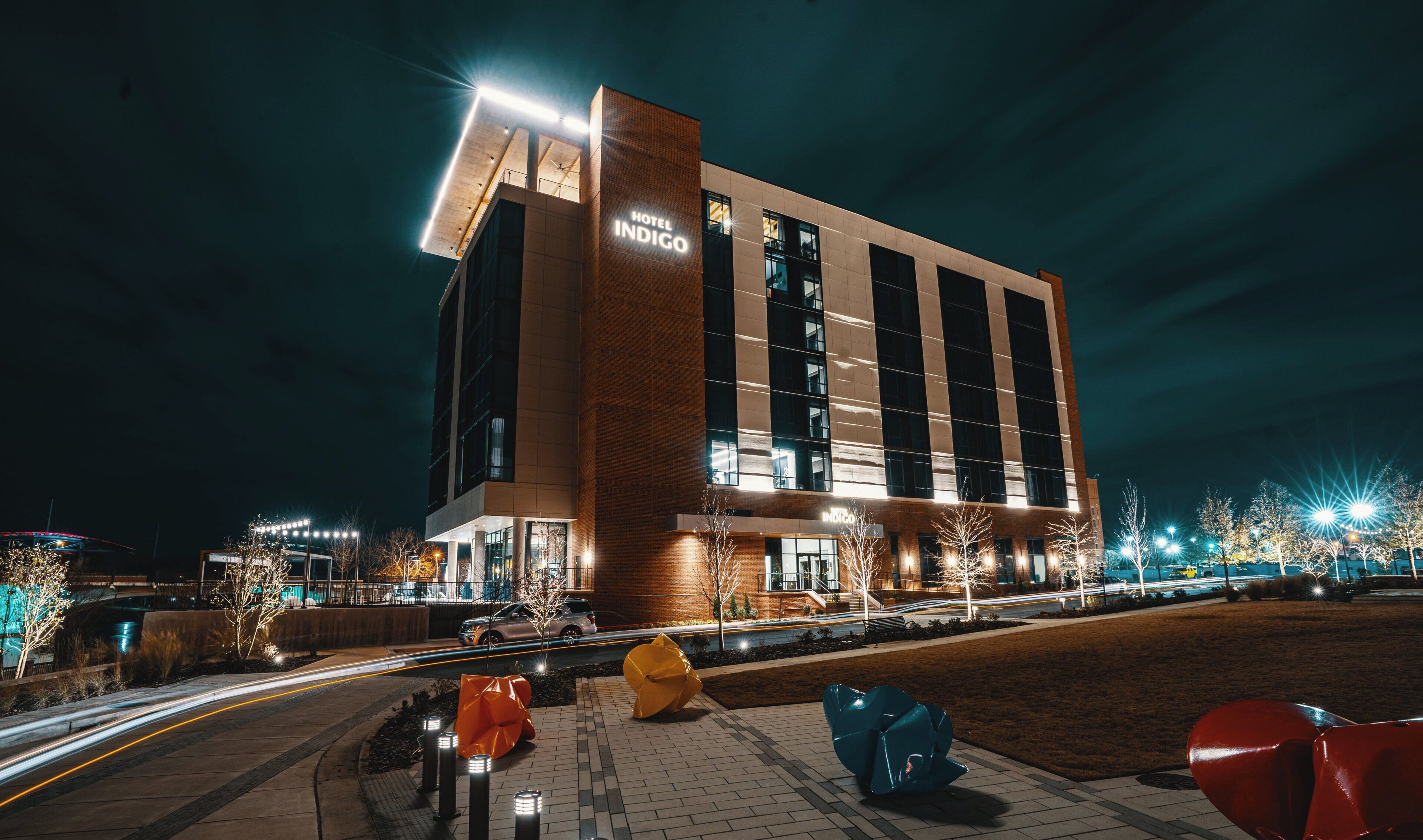 A modern hotel building with a prominent sign, a well-lit entrance, and a parking area with a distinctive sculpture.