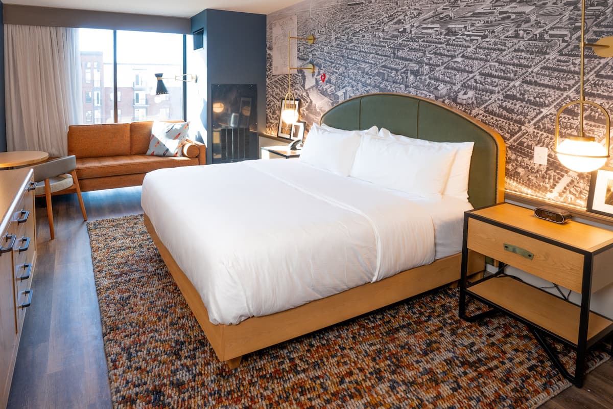 A hotel room with a large bed, a sofa, a desk, and a cityscape mural on the wall.