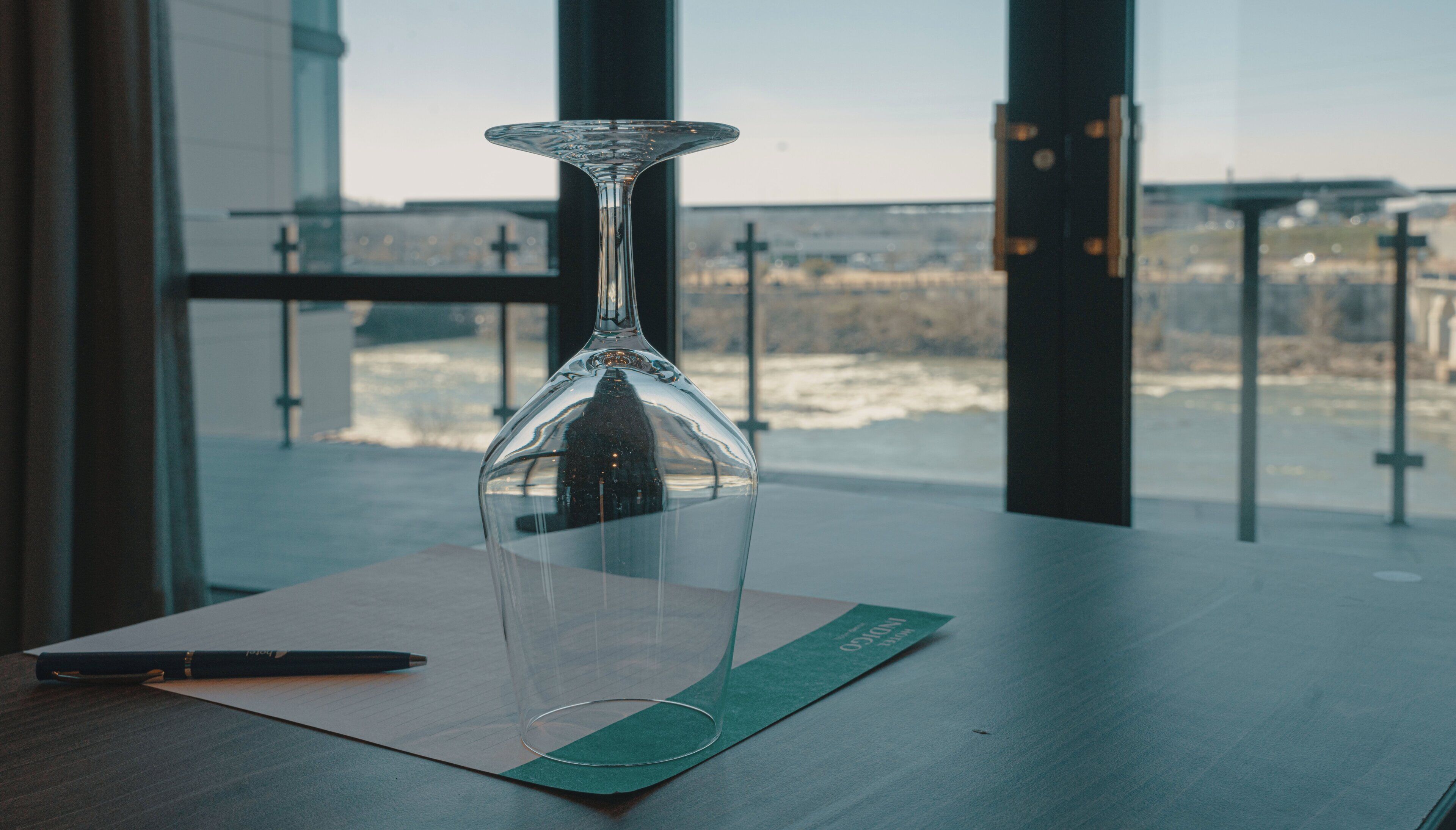 A clear glass hourglass on a wooden table with a pen beside it, set against a window with a view of a bridge and buildings.