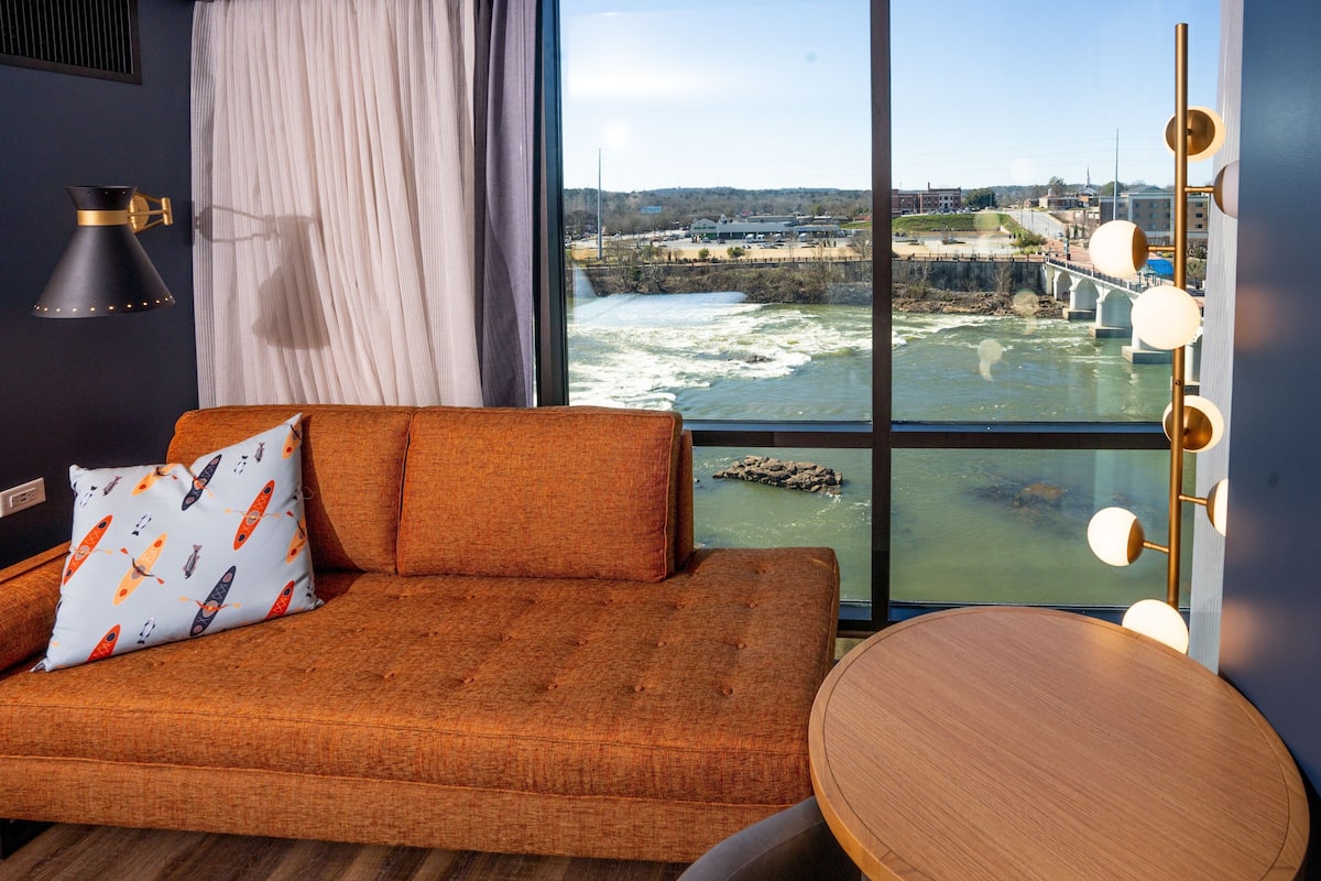 A room with a large window offering a view of a river and bridge, a sofa with patterned pillows, and a round wooden table.