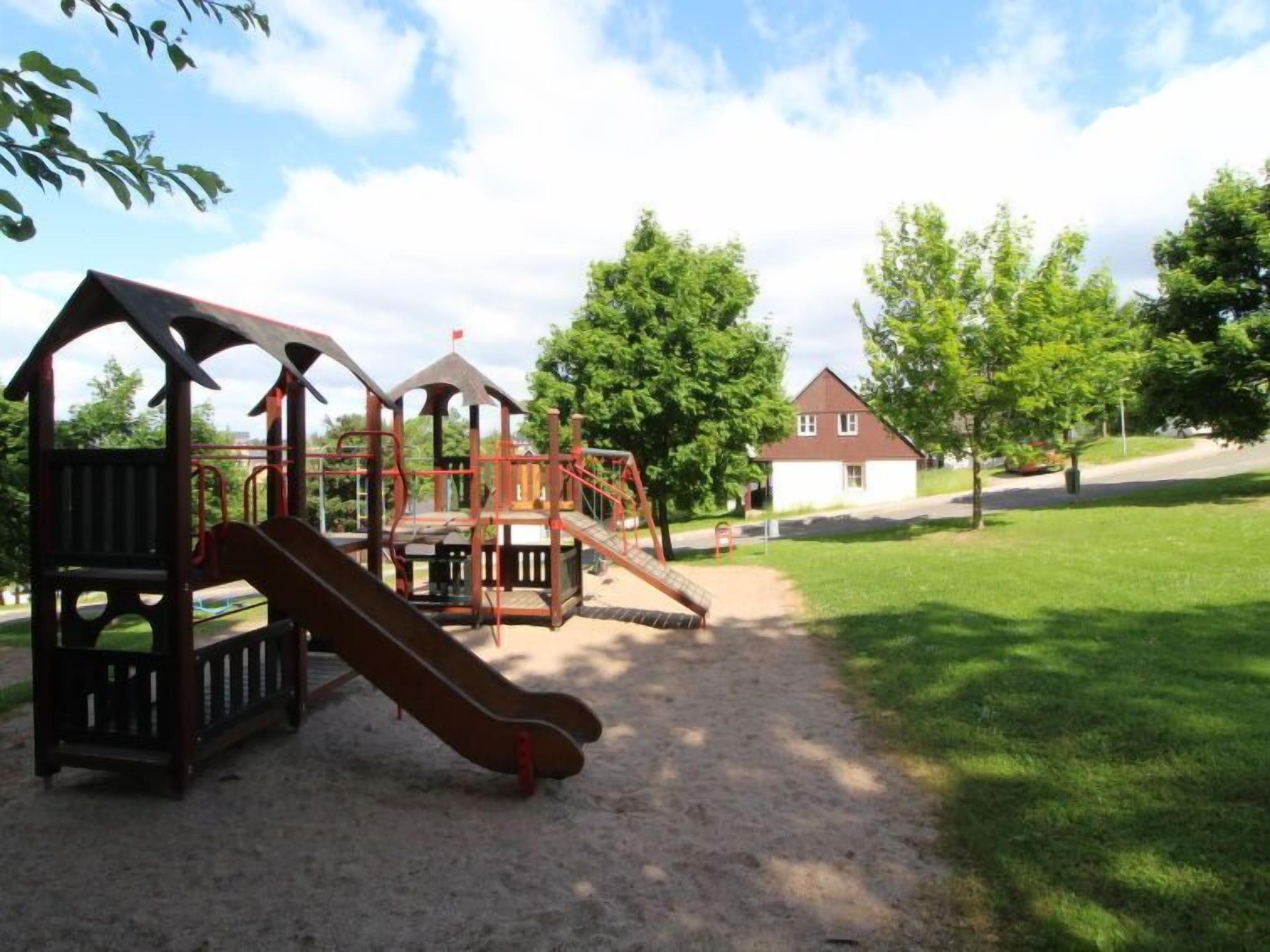 Ein Spielplatz mit Rutsche und Klettergerüsten, umgeben von Bäumen und einem Haus im Hintergrund.