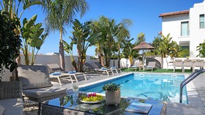 A poolside area with lounge chairs, a glass table with a fruit bowl, and a view of a building in the background. - VILLA APOLLONIA - Brand new villa walking to Trinity Beach (Paralimni)