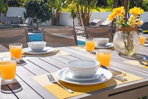 A dining table set with white bowls, plates, and cutlery, accompanied by glasses of orange juice, in an outdoor setting with a pool and lounge chairs in the background. - VILLA APOLLONIA - Brand new villa walking to Trinity Beach (Paralimni)