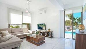 A modern living room with a sofa, coffee table, TV, and a view of a pool. - VILLA APOLLONIA - Brand new villa walking to Trinity Beach (Paralimni)