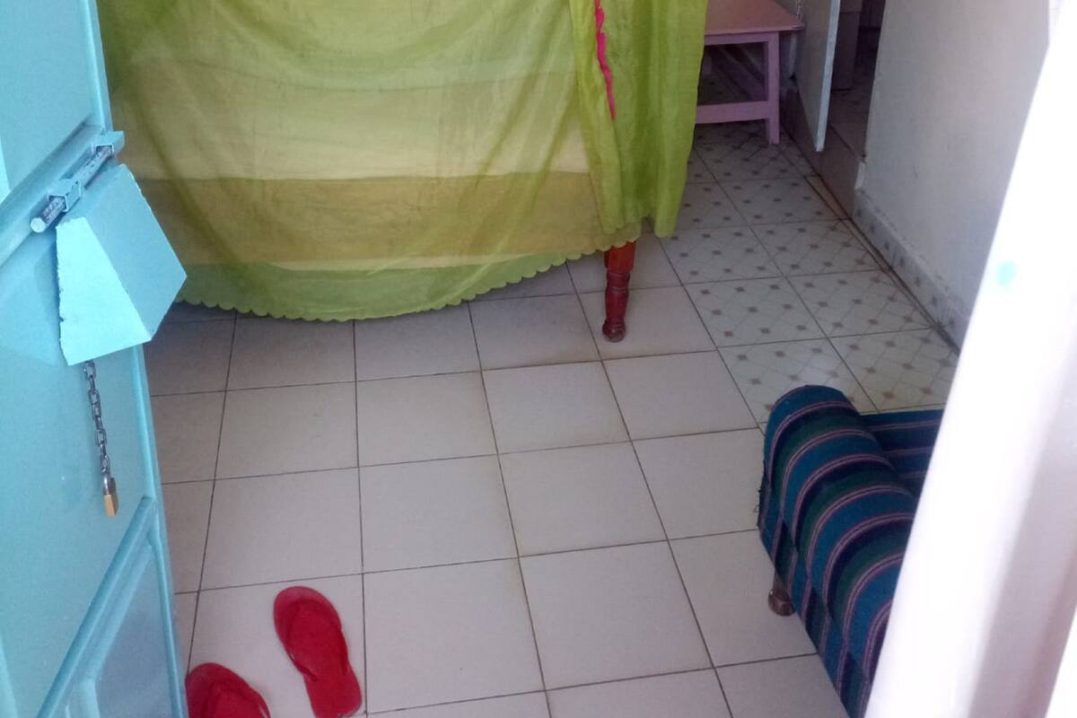 Quarto com uma cama coberta por um dossel verde, um pequeno banquinho de madeira e um cobertor listrado no chão.