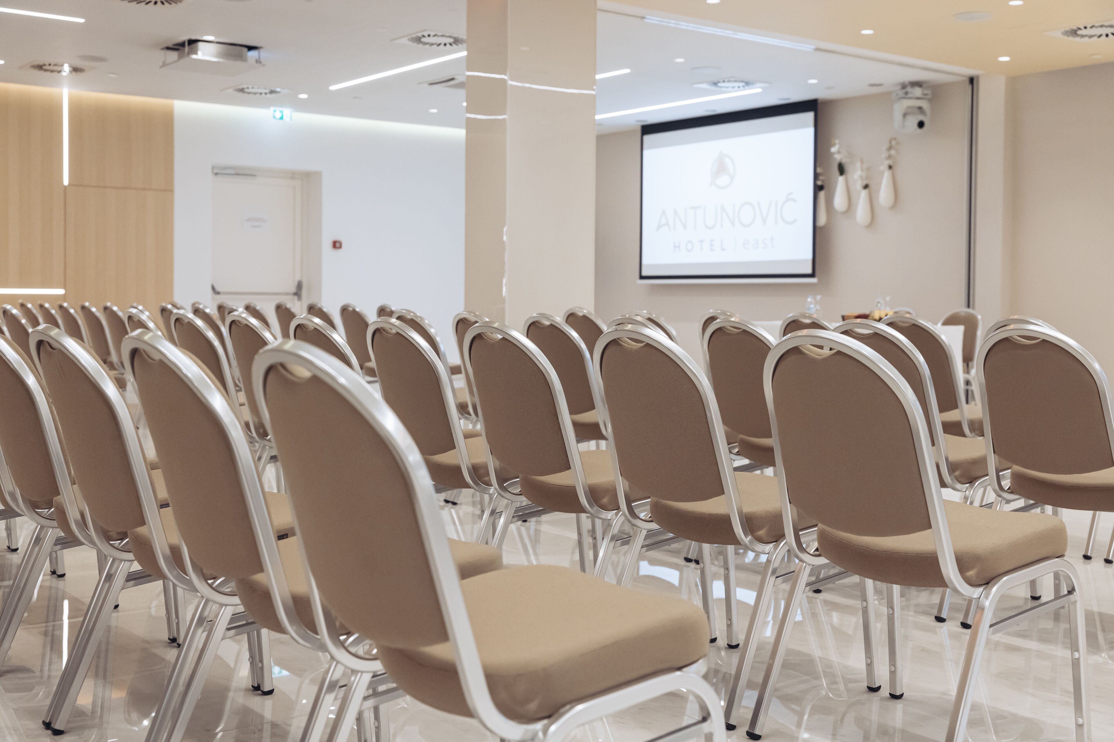 Uma sala de conferências com fileiras de cadeiras bege, uma tela de projeção e um pódio.