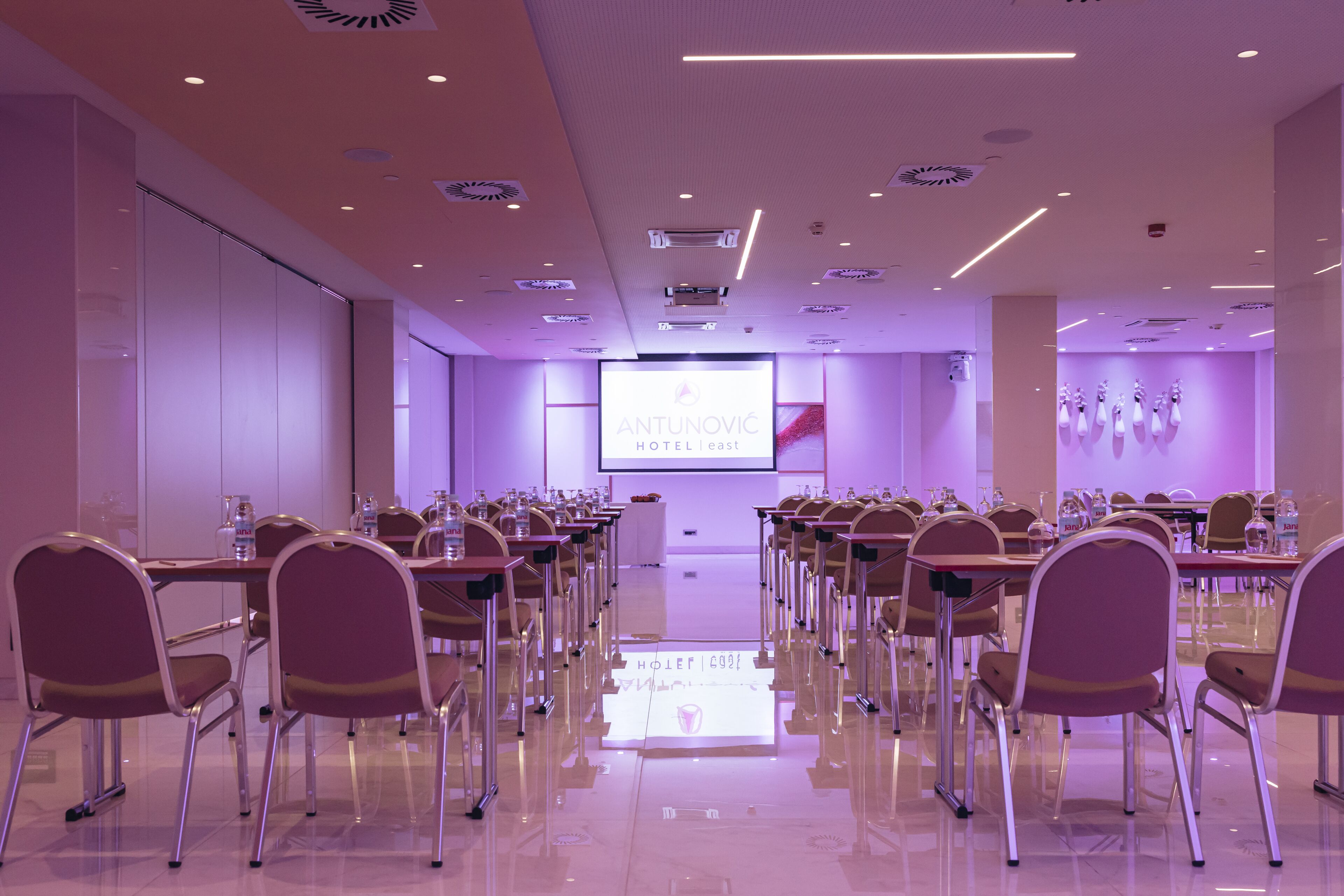 Un salón de conferencias con filas de sillas, una pantalla grande al frente y un diseño moderno del techo.