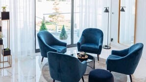 Een moderne woonkamer met een ronde salontafel, vier blauwe fauteuils en uitzicht op een tuin door grote ramen.