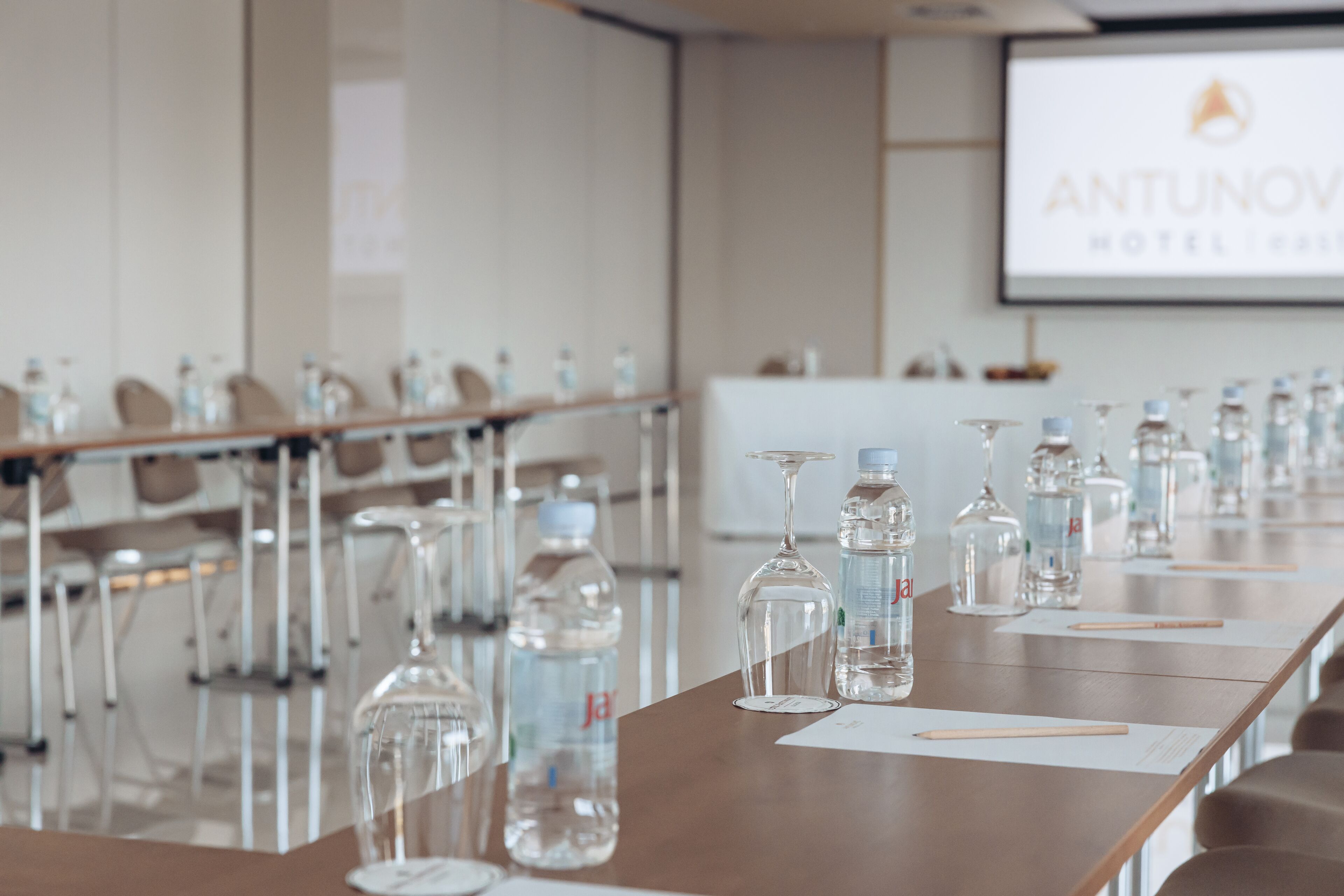 A conference room with a long table, water bottles, and glasses. A screen displays the Antunov Hotel logo.