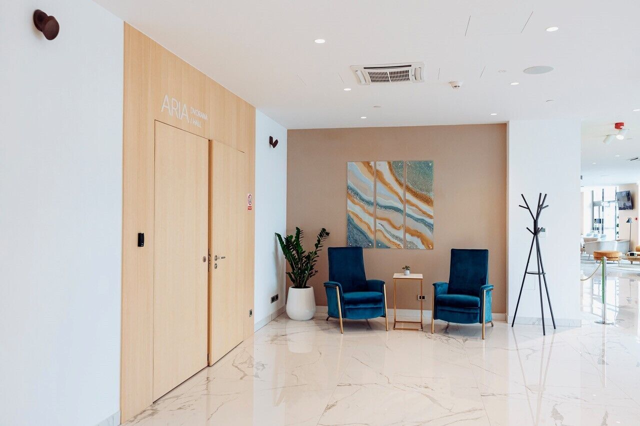 Una moderna zona de recepción con una puerta de madera que dice "ARIA", suelo de mármol, dos sillones azules, una mesita y un cuadro abstracto grande, además de una lámpara de pie.