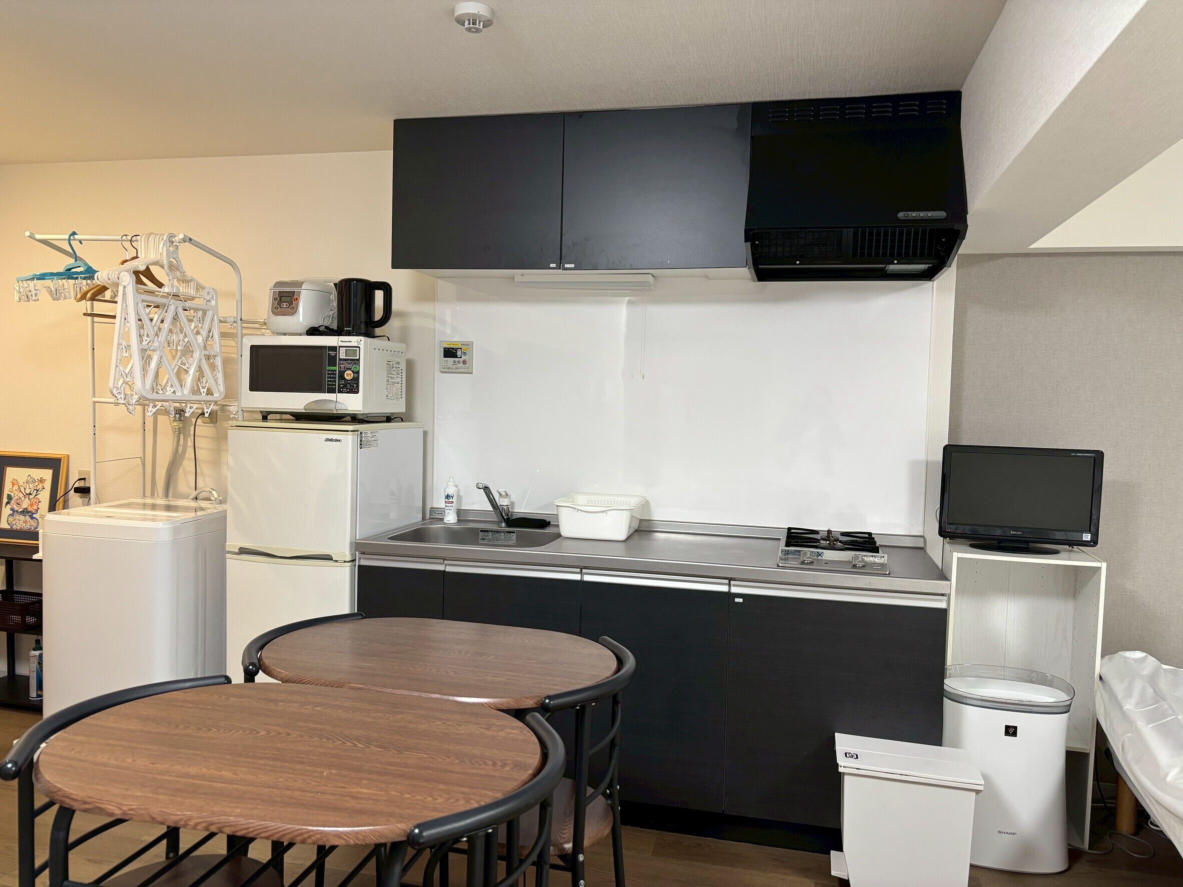 A compact kitchenette with a microwave, coffee maker, and sink. There are two wooden chairs and a small trash bin.
