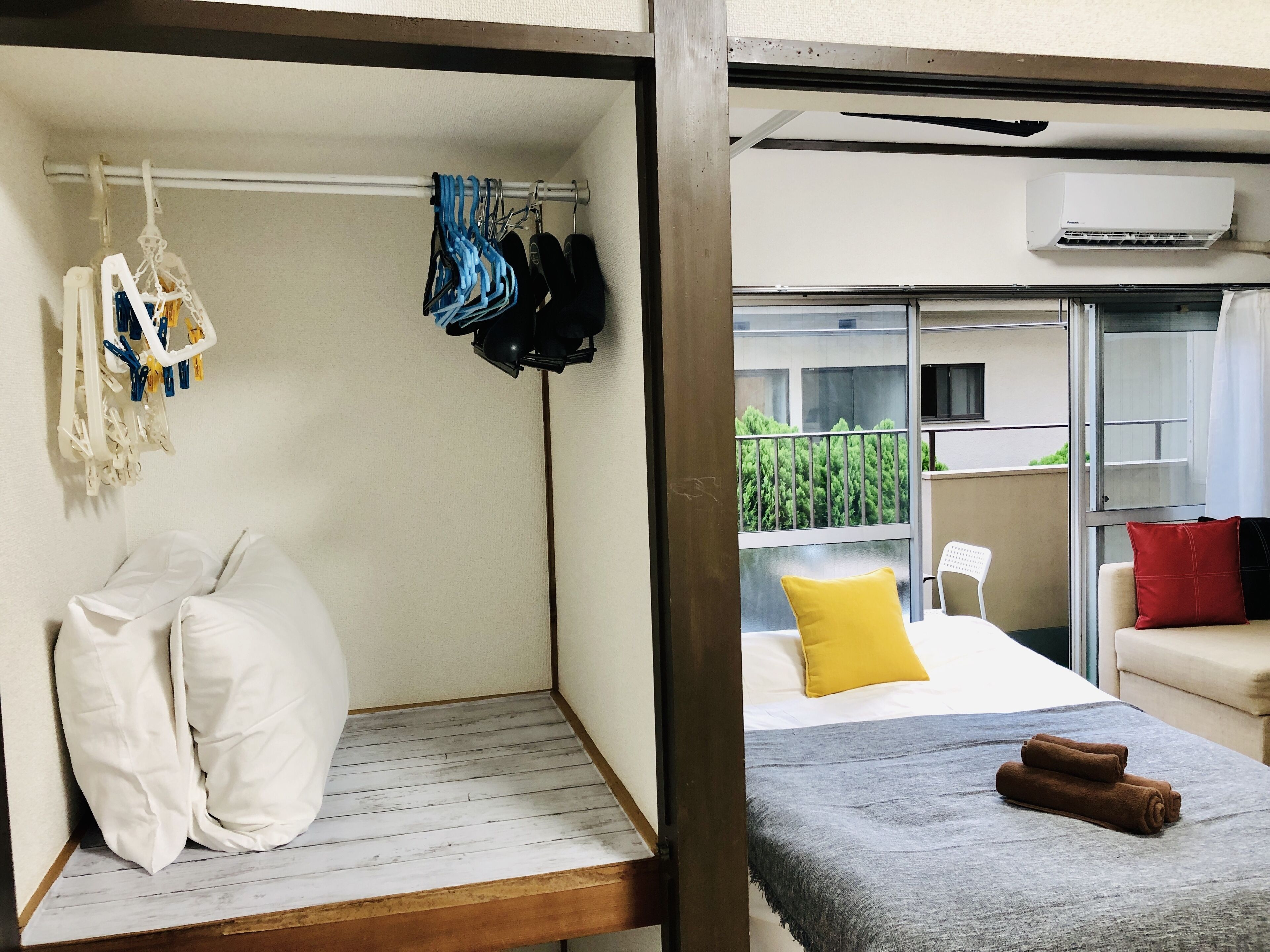A compact room with a bed, a small seating area, a window with a view of greenery, and a clothes rack with various items.