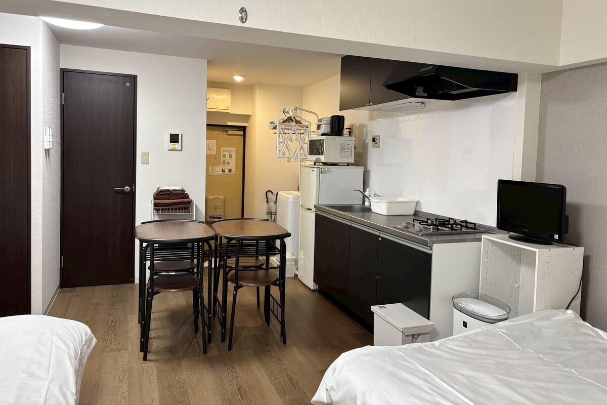 A compact hotel room with a kitchenette, a dining area with two chairs, a bed, and a small white cabinet.
