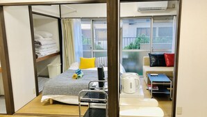 A compact hotel room with a bed, a small table with a kettle, a sofa, and a view of the outdoors through large windows.