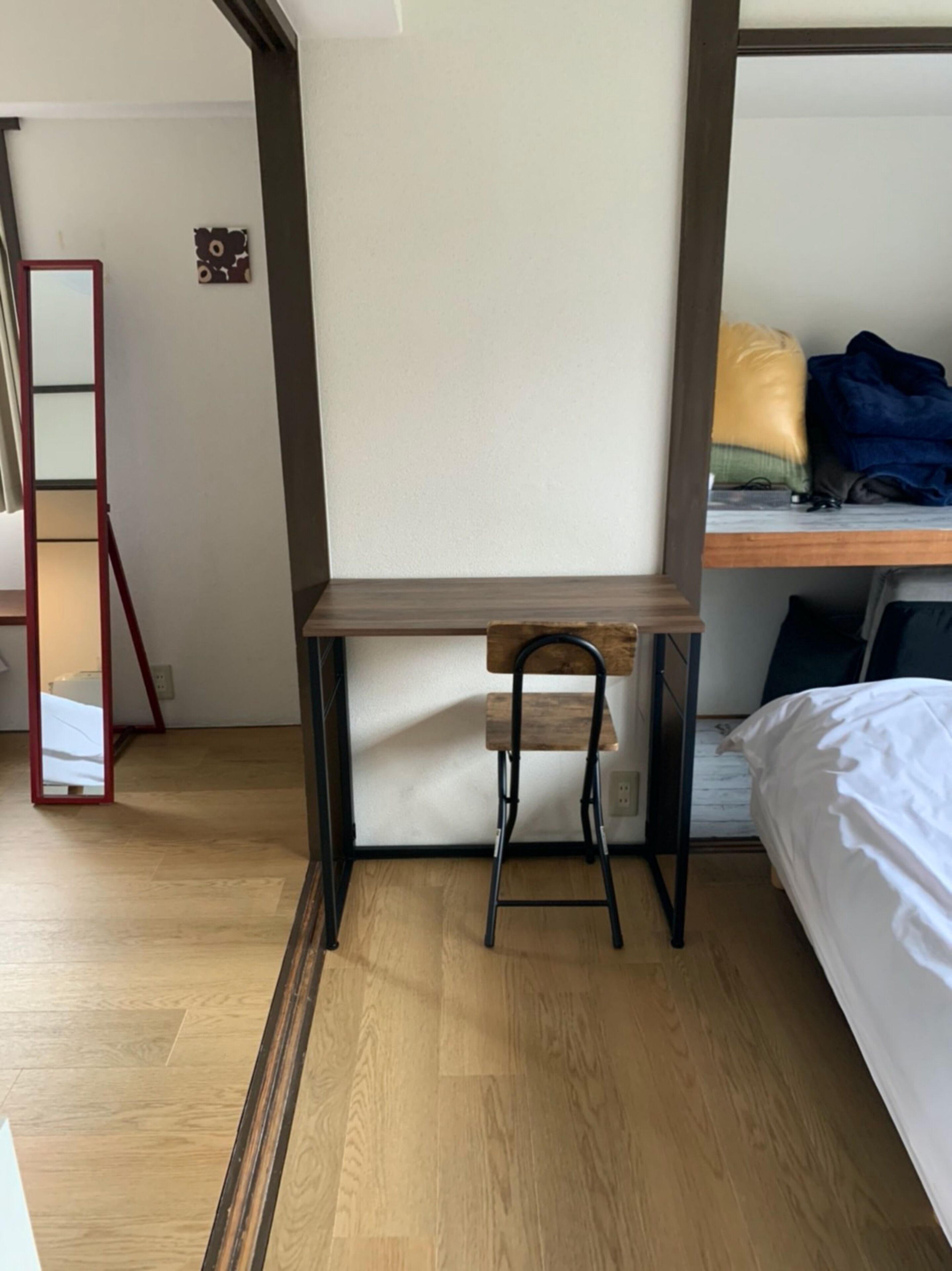 A compact room with a wooden desk, a chair, a bunk bed with a mattress and pillows, a small mirror, and a wooden floor.
