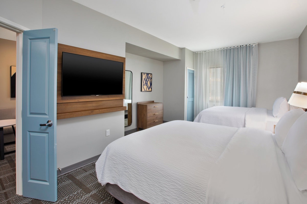 A hotel room with two beds, a TV mounted on the wall, a wooden dresser, and a window with curtains.