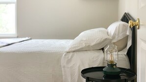 A neatly made bed with white bedding, a bedside table with a lamp, and a window with a view of greenery.