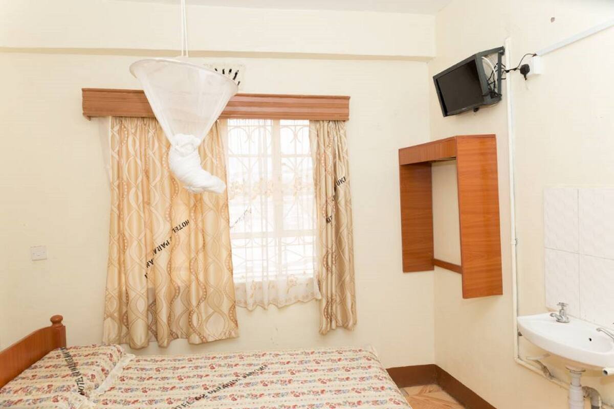 Un dormitorio con cama, ventana con cortinas, mesita de noche, televisor montado en la pared y baño con lavamanos y espejo.
