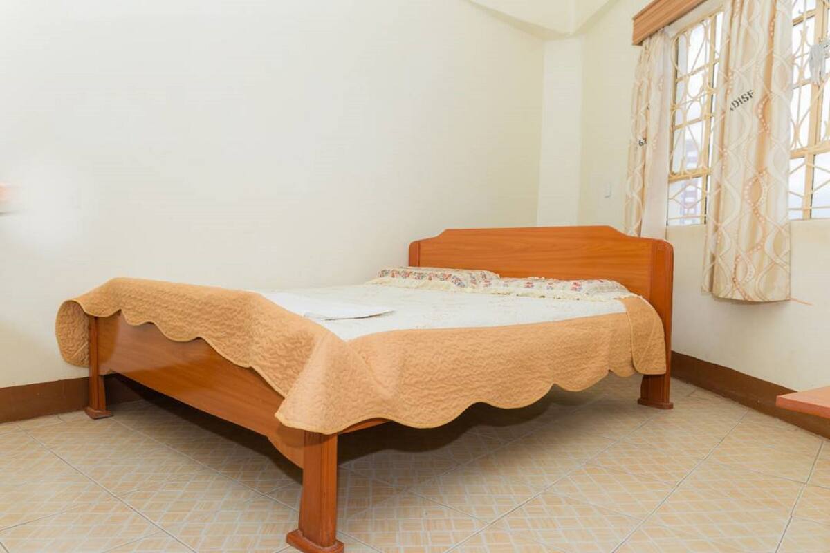 Una cama individual con estructura de madera y colcha color beige en una habitación con suelo de baldosas y una ventana con cortinas.