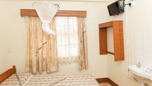 A bedroom with a bed, a window with curtains, a bedside table, a mounted television, and a bathroom with a sink. - Sixty Two Paradise Hotel (Nanyuki)