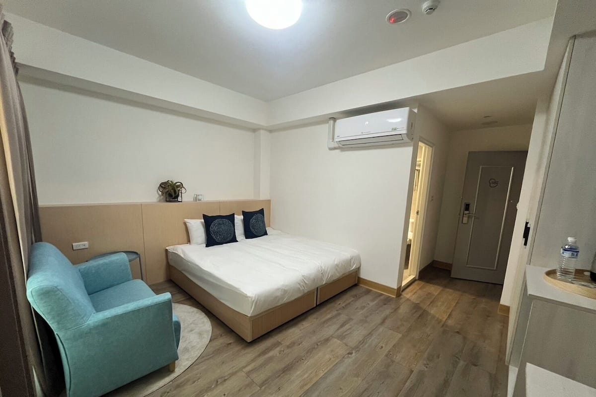 A bedroom with a bed, a blue chair, a small table, and an air conditioning unit.