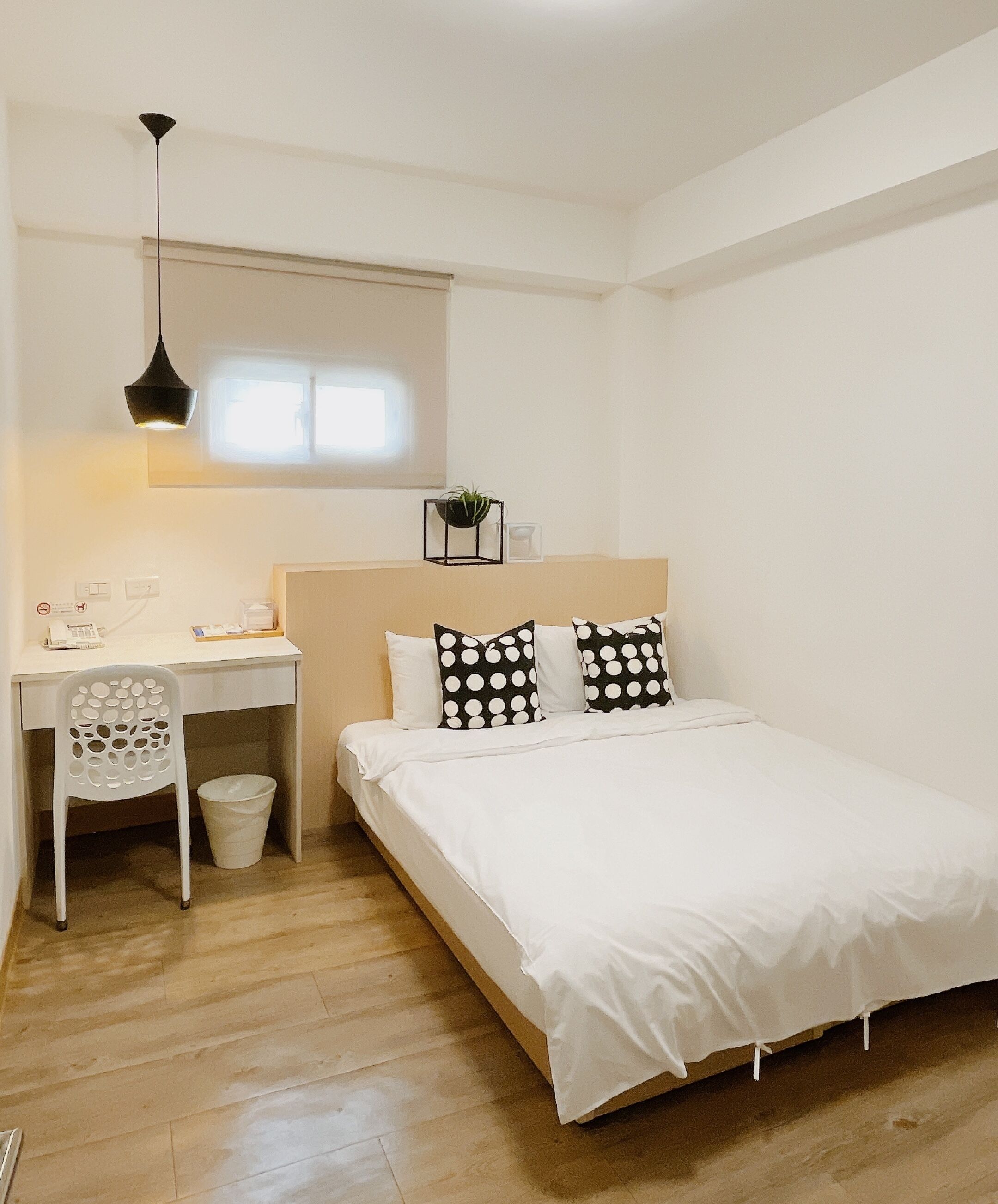 A bedroom with a bed, a desk, a chair, a hanging light fixture, and a small plant on the wall.