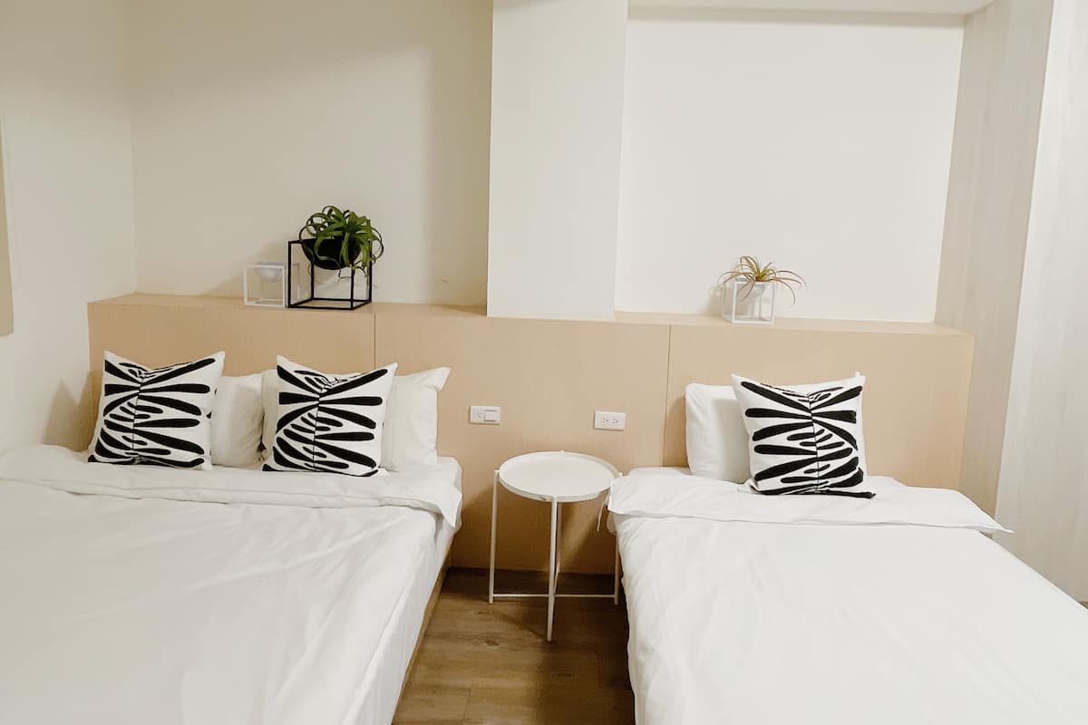 A hotel room with two beds, white bedding, and black patterned pillows. There is a small round table and two potted plants on the headboard.