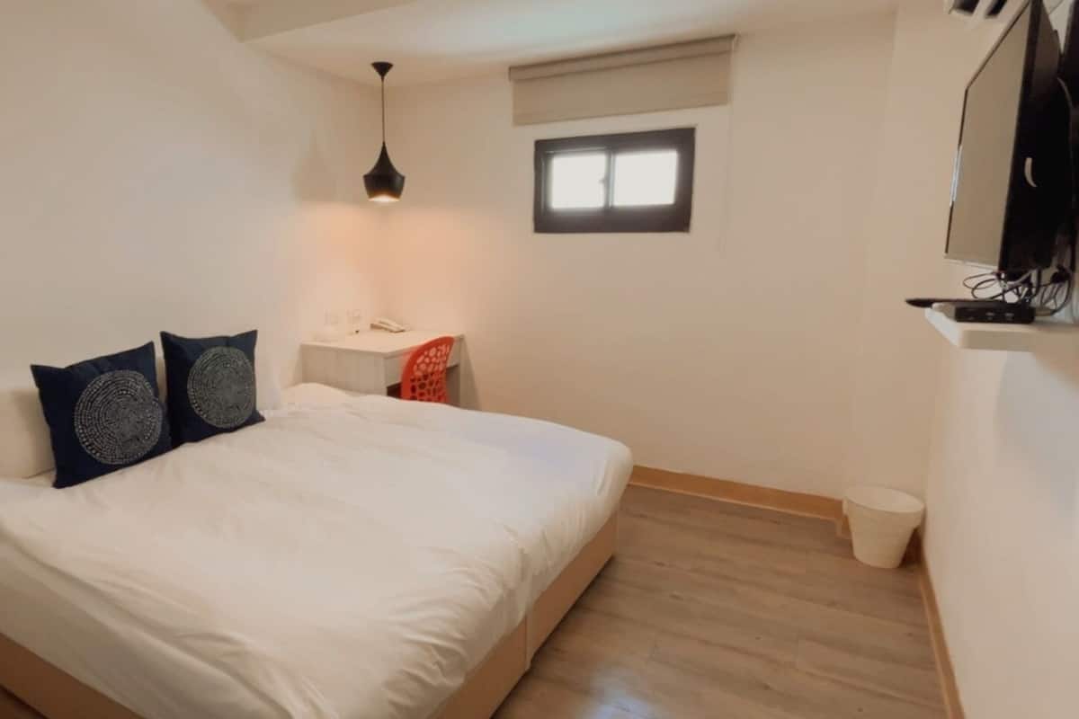 A bedroom with a bed, a desk, a chair, a small table, a window, a ceiling light, and a TV mounted on the wall.