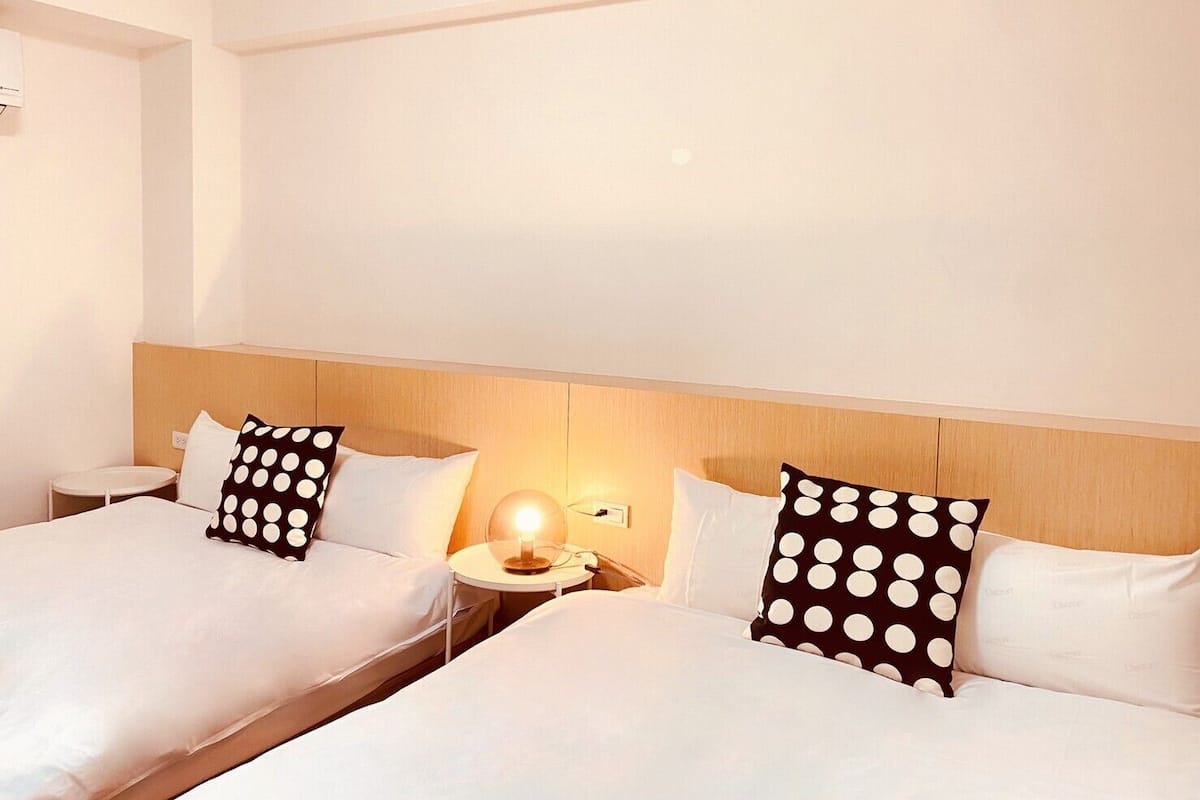 A hotel room with two beds, each with white bedding and black pillows with white polka dots. There is a bedside table with a lamp between the beds.