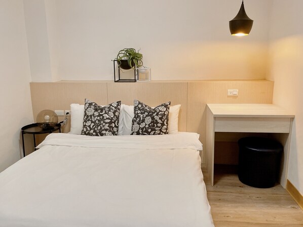 Habitación de hotel ordenada con cama, escritorio, una planta pequeña y un cesto de basura.