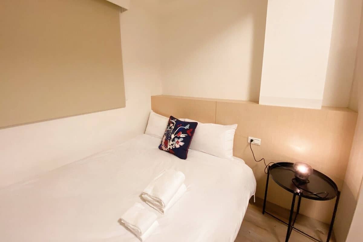 A hotel room with a bed, a pillow, a folded towel, and a small round table with a lamp.