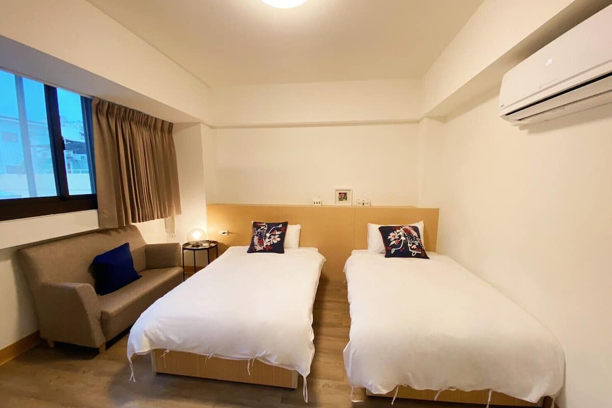 A hotel room with two single beds, a sofa, a small table, and an air conditioning unit.