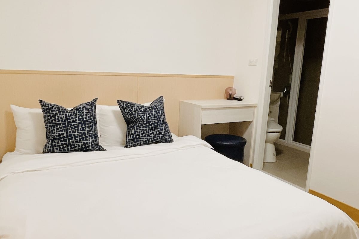 A hotel room with a large bed, two patterned pillows, a bedside table with a lamp, and a bathroom visible through an open door.