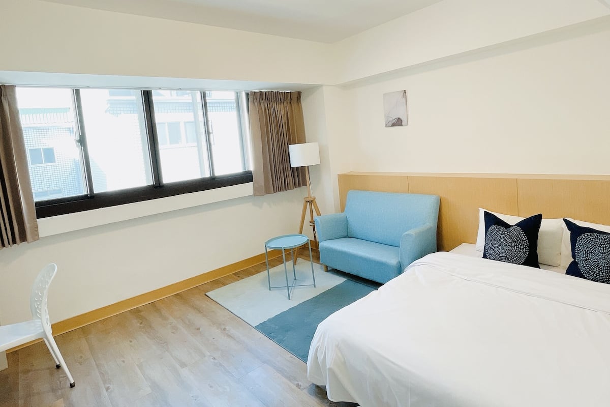 A hotel room with a bed, a blue sofa, a white chair, a small table, and a lamp.