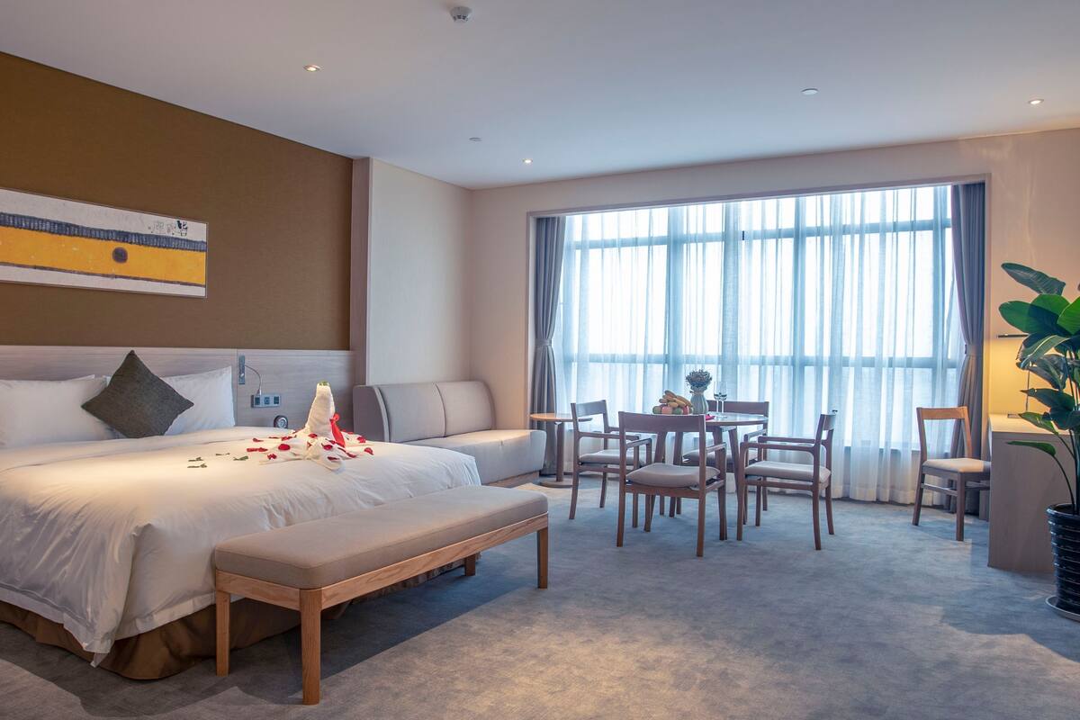 A modern hotel room with a large bed, a sofa, a dining table with chairs, and a large window with curtains.