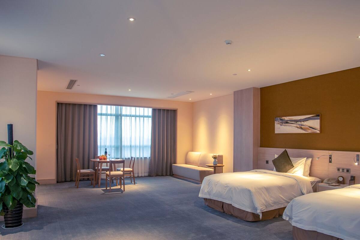 A hotel room with two beds, a dining area with a table and chairs, a large window with curtains, and a potted plant.
