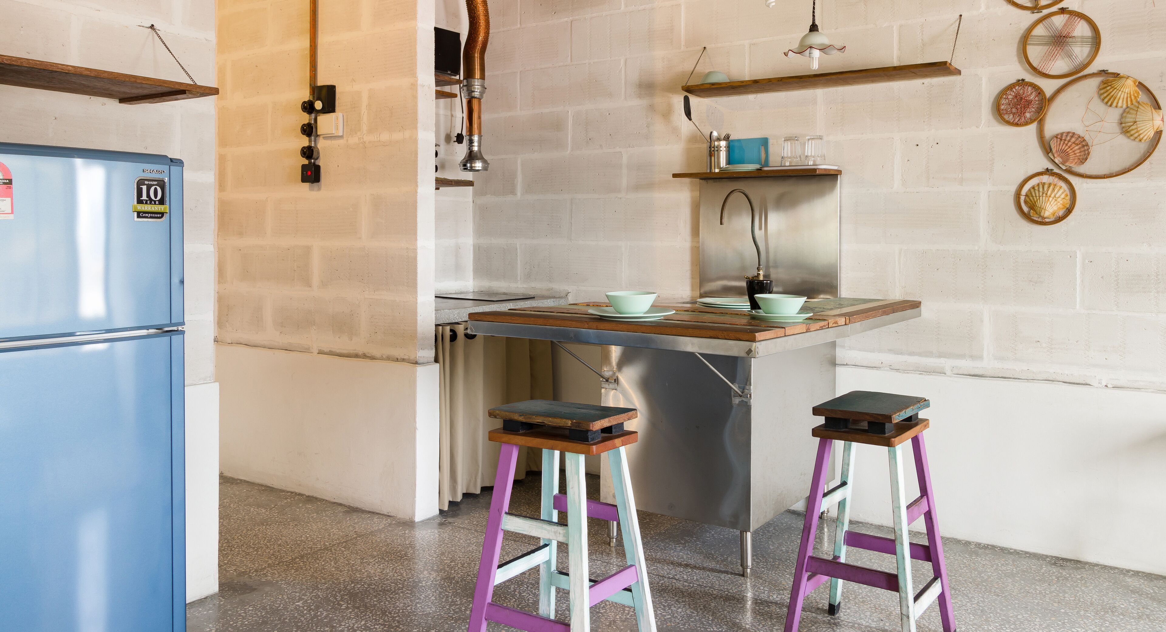 A compact kitchen with a blue refrigerator, a stainless steel sink, and a wooden countertop. There are two stools and a shelf with decorative items.