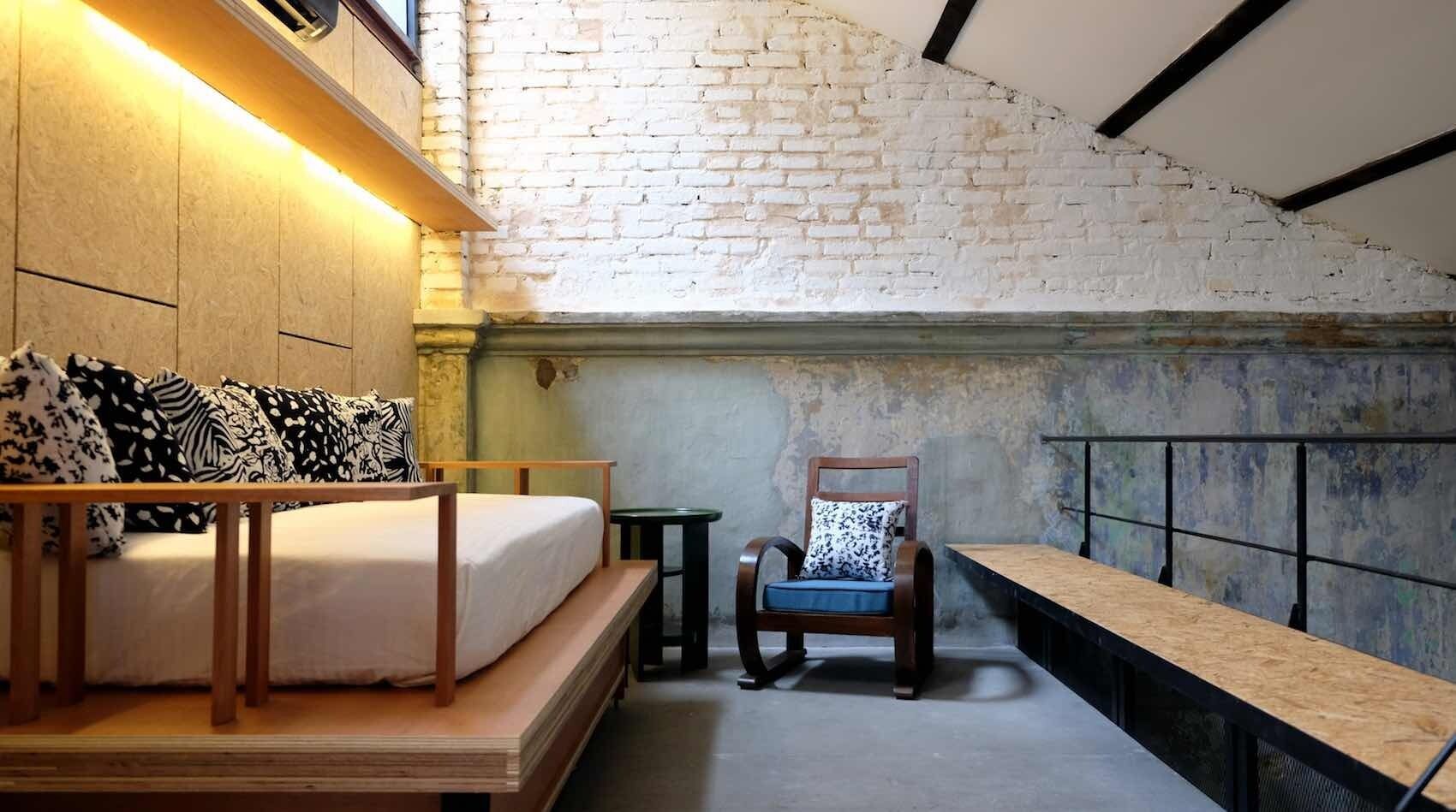 A loft-style bedroom with a bed, a chair, and a small table. The room has exposed brick walls and a sloped ceiling.