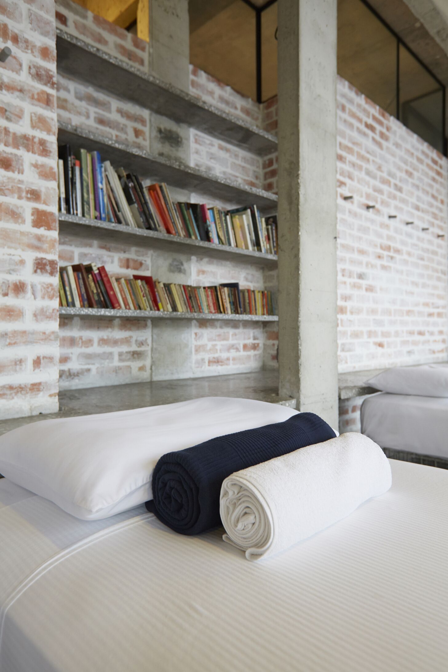 A hotel room with a brick wall, a bookshelf filled with books, a neatly made bed with a rolled towel, and a white pillow.