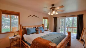 A bedroom with a wooden bed, a nightstand, a ceiling fan, and a view of the outdoors through a window. - Wraparound Deck, Stunning Mountain Views, Near Downtown Pagosa & Wolf Creek Ski Area (Pagosa Springs)