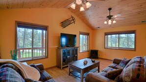 A cozy living room with a television, wooden furniture, and large windows offering a view of the outdoors. - Wraparound Deck, Stunning Mountain Views, Near Downtown Pagosa & Wolf Creek Ski Area (Pagosa Springs)