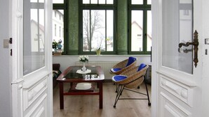 Une pièce avec une table à plateau de verre, des chaises en osier, un vase de fleurs et une vue dégagée sur l’extérieur grâce à de grandes fenêtres.