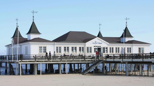 Un bâtiment blanc doté de trois tours surmontées de toits verts, d’une jetée en bois et d’un escalier menant à l’entrée.