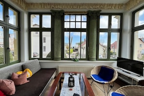 A room with a sofa, a table with a bottle, a chair, a TV, and large windows with curtains.