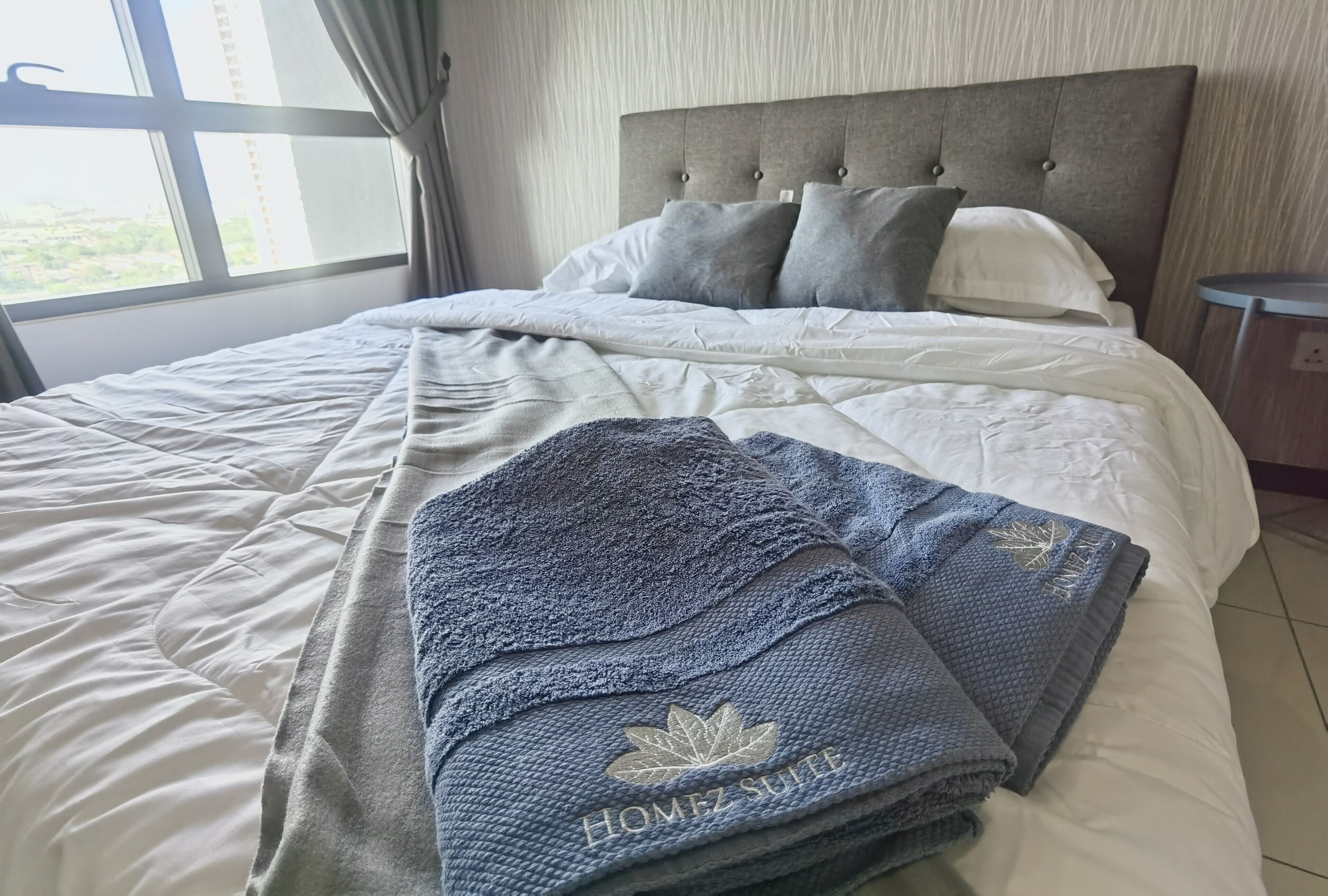A neatly made bed with a grey and white duvet, a grey blanket with a logo, and a grey headboard.