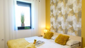 A neatly made bed with yellow pillows and a rolled towel, a bedside table with a lamp, and a window with a plant. - Apartments Banonia - Morning Dawn (Verzej)