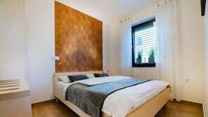Ein Schlafzimmer mit einem hölzernen Kopfende, einem Bett mit weißer Bettwäsche, einem Fenster mit Jalousien und einem kleinen Blumentopf auf der Fensterbank.
