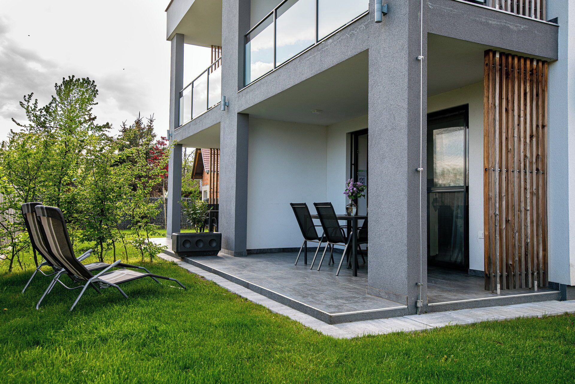 A modern two-story building with a covered outdoor area featuring a table and chairs, a concrete planter, and a wooden fence.
