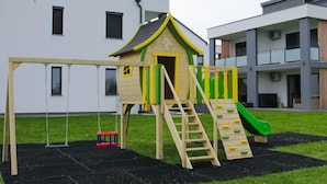 Een speelplaats met een glijbaan en schommels, gelegen voor een modern gebouw met balkons.